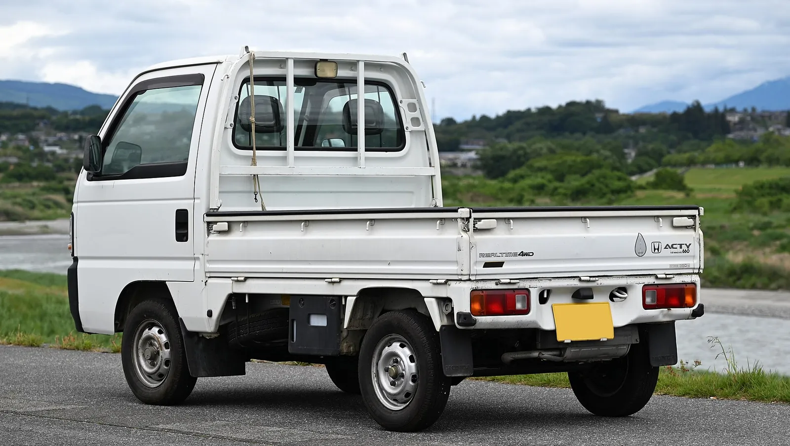 White Honda Acty SDX 4WD kei truck parked by a river with green hills behind