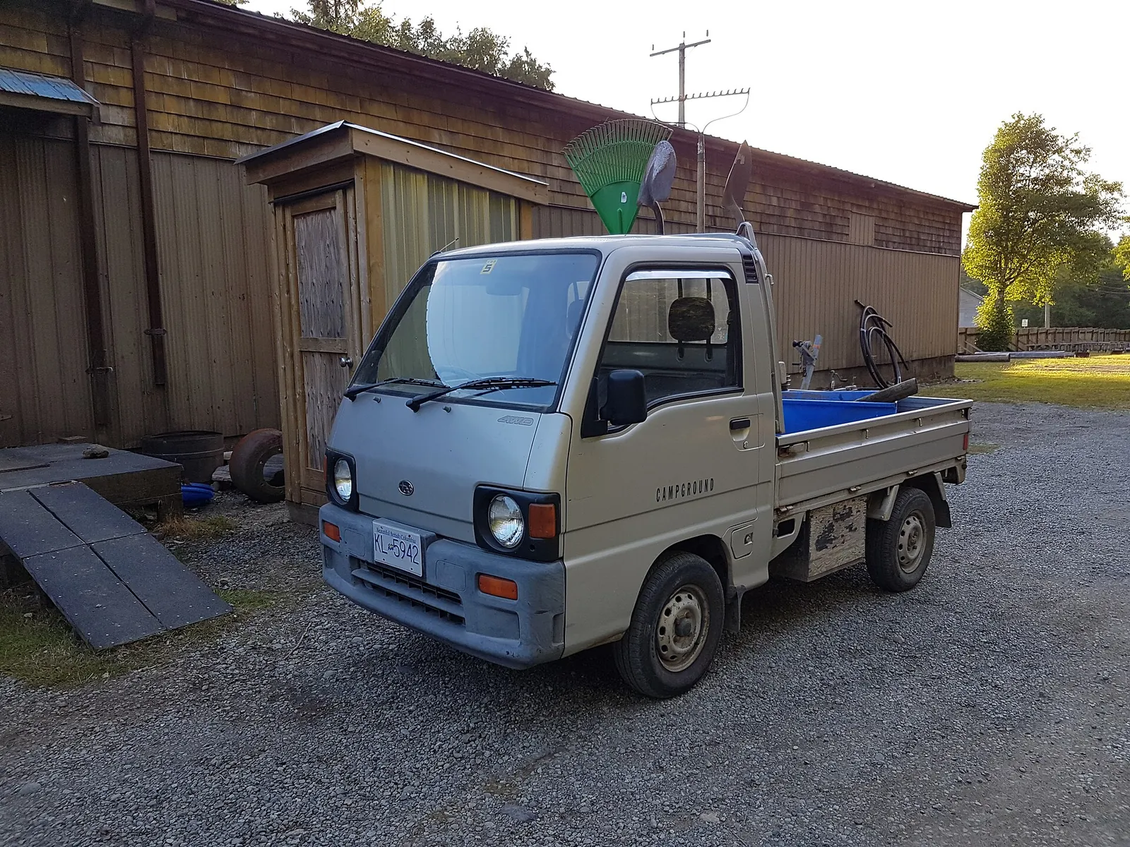 Subaru Sambar kei truck with tools in the bed, parked on gravel by a wooden shed at a campground