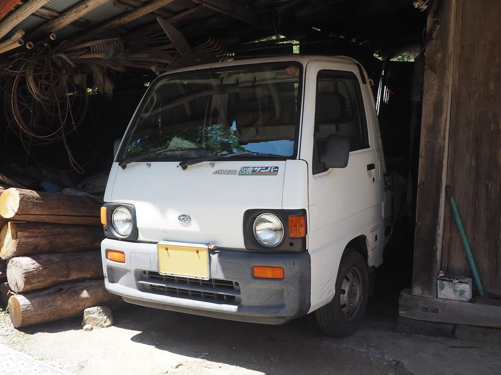White Subaru Sambar 4WD farming truck parked in a Japanese wooden shed beside stacked logs