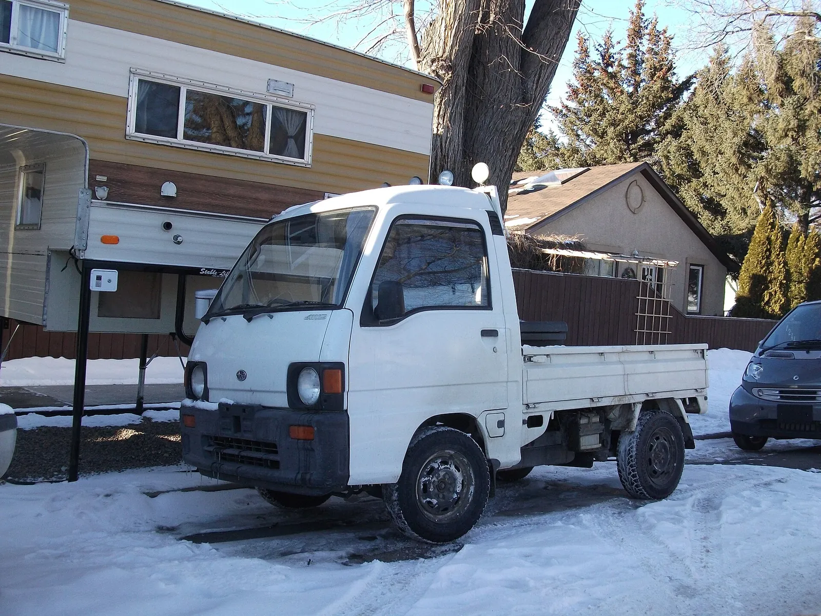 White Subaru Sambar kei truck parked in snow beside a house and a camper